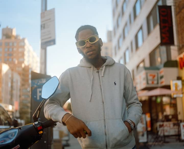 A man in sunglasses poses on a New York City street with a motorcycle and urban backdrop.
