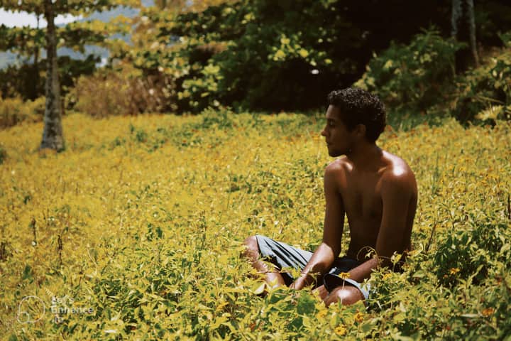 Shirtless man sitting in a sunny meadow surrounded by wildflowers.