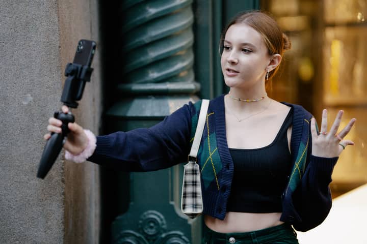 Young woman in casual attire takes a selfie outdoors using a smartphone and stand.