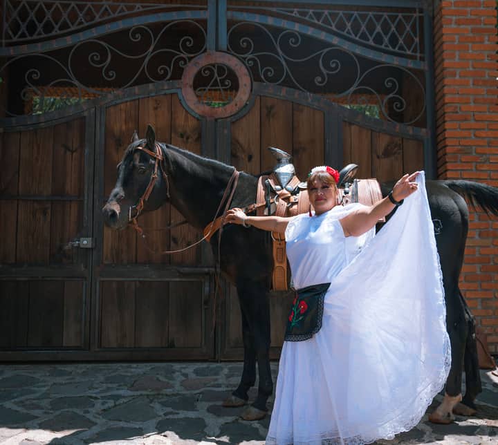 A woman in a white dress and traditional attire poses joyfully beside a black horse.