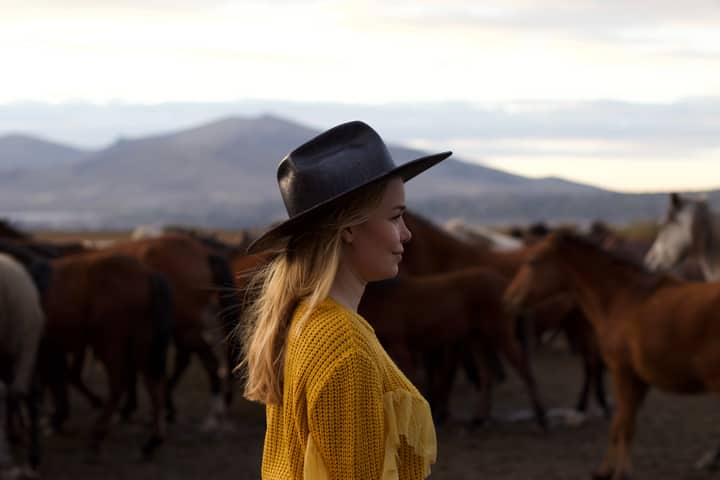 A woman in a cowboy hat and yellow top stands with horses in a scenic countryside setting.