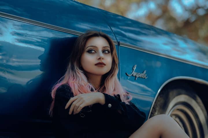 A young woman with pink hair leans against a vintage blue Impala car, with a thoughtful expression.