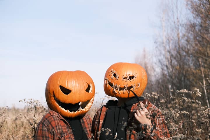Two people wearing pumpkin heads in a field during fall, creating a spooky Halloween scene.