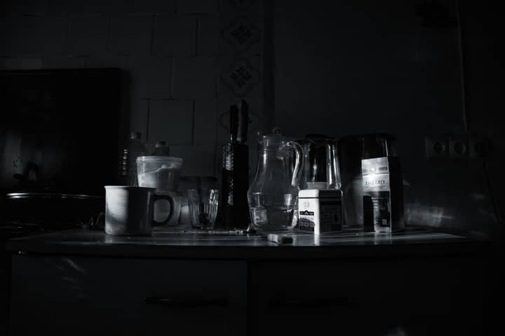 A moody kitchen scene featuring glassware, mugs, and containers in monochrome lighting.
