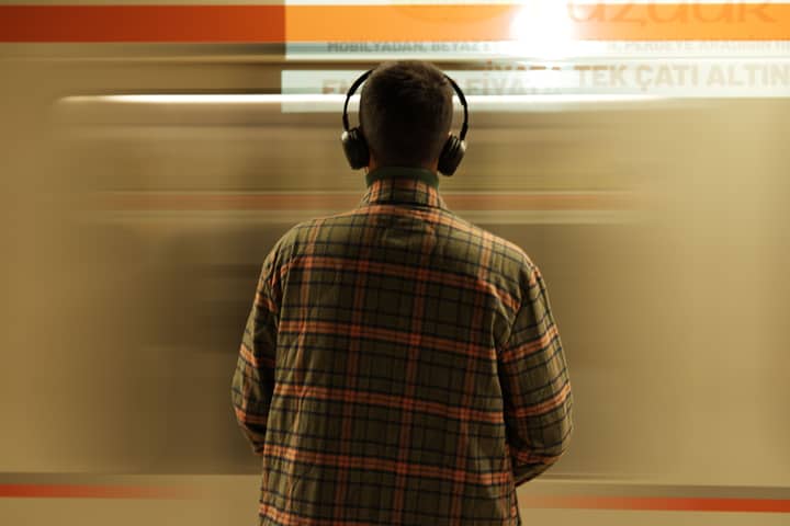 A man wearing headphones stands at a subway station, observing a passing train.