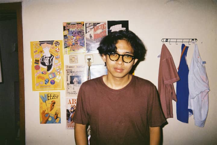 A young adult with glasses in a room with vibrant posters on the wall.