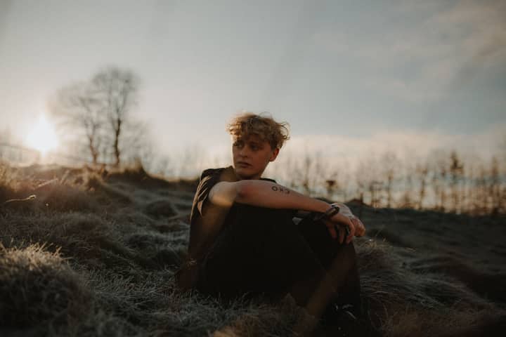 Young man sitting alone in a grassy field during sunset, conveying a sense of solitude and relaxation.