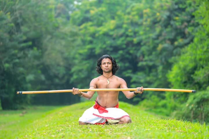A shirtless Asian man practicing Silambam with a stick on a grassy field.