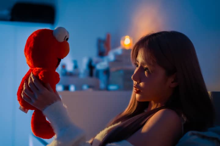 A woman with dyed hair poses holding a plush toy in a softly lit room.