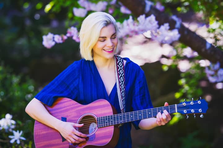 Blonde woman in blue dress playing guitar in a vibrant spring garden with blossoms.