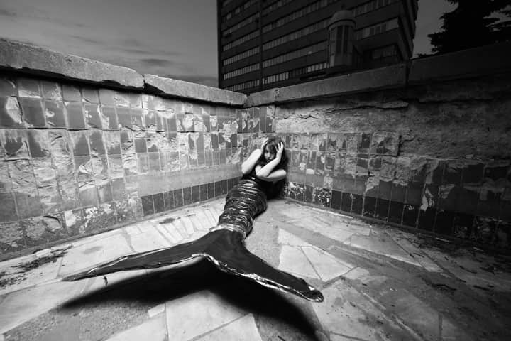 Black and white photo of a merperson posing in an urban corner, evoking mystery and fantasy.