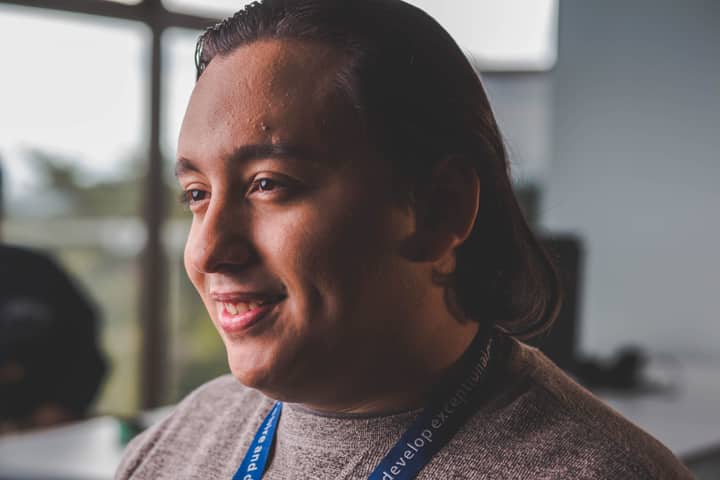Portrait of a smiling man with long hair, wearing a sweater, in a bright office setting.