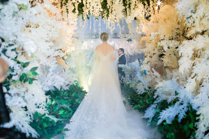 Stunning bride in wedding dress walking down a floral aisle, surrounded by lush greenery and white flowers.