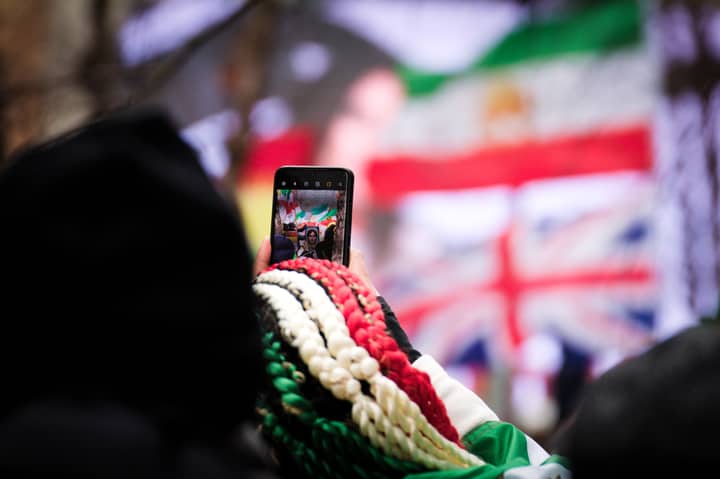 Person using smartphone to capture live event in Brussels with visible flags in background.