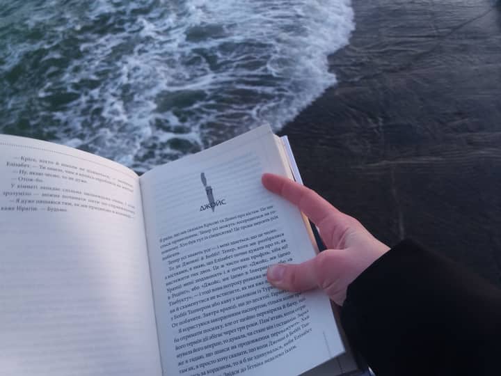 A person holds and reads a book by the ocean, enjoying a peaceful moment by the water.