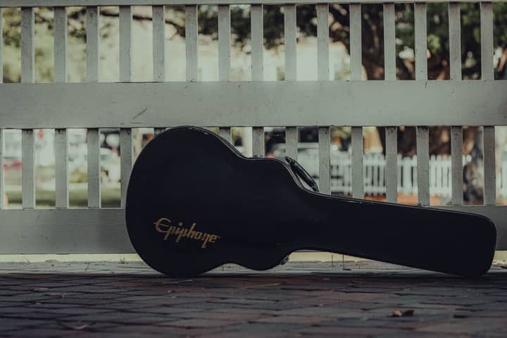 A guitar case rests against a white fence on a pavement outdoors.
