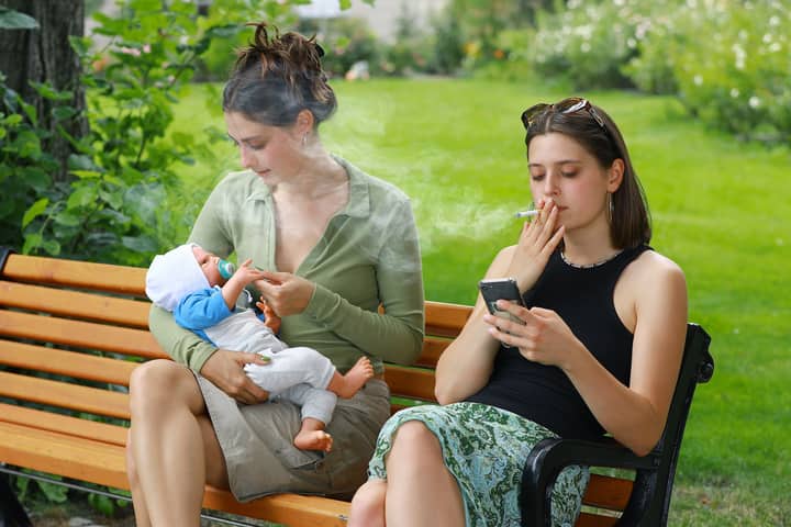 Two women sitting on a park bench, one with a baby, the other smoking a cigarette and using a smartphone.