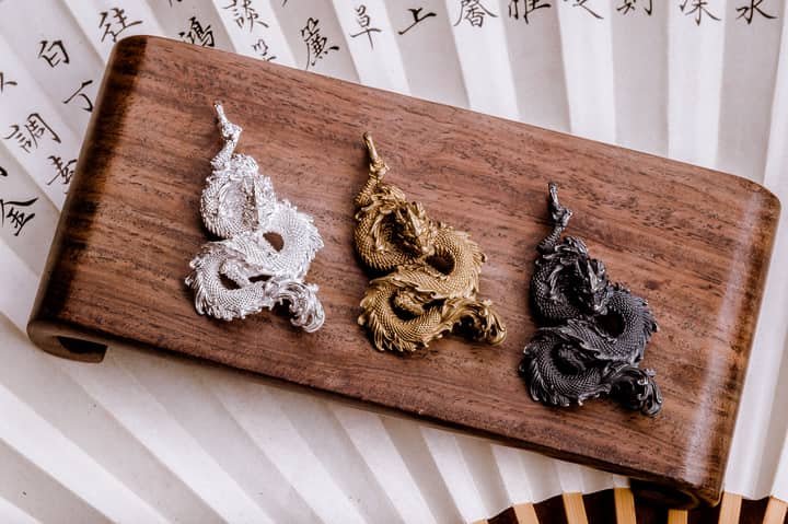 Three intricately designed dragon pendants in silver, gold, and black on a wooden stand.