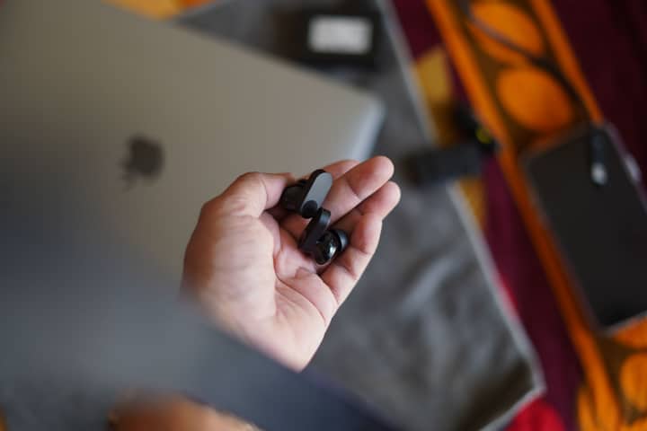 Hand holding black wireless earbuds with tech gadgets in the background, perfect for tech lifestyle concepts.