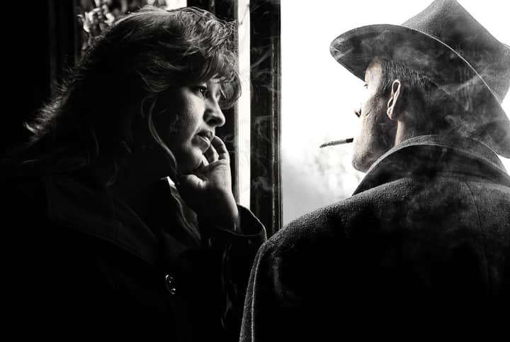 A contemplative couple in a black and white photo, with a cigarette and window adding to the mood.