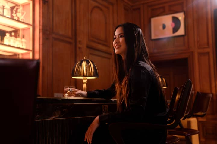 Woman with long hair sipping whiskey at a stylish bar in warm lighting, capturing elegance and relaxation.