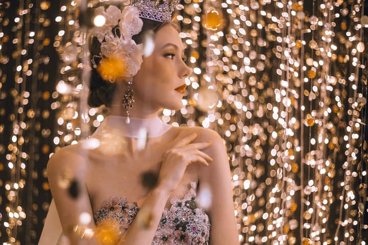A woman wearing a crown and floral dress elegantly poses amidst sparkling string lights.