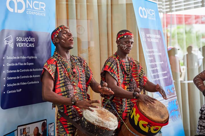 Two African drummers in traditional attire performing at a cultural festival indoors.