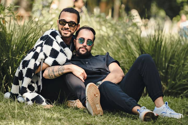 Two men wearing sunglasses, enjoying a relaxed moment on a grassy field with greenery in the background.