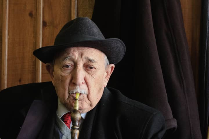 Senior man in a suit and hat smoking shisha indoors, symbolizing leisure and retirement.