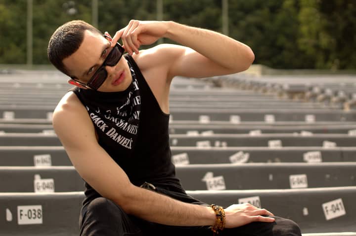 Fashionable young man in black tank top sitting confidently on empty bleachers, adjusting sunglasses.