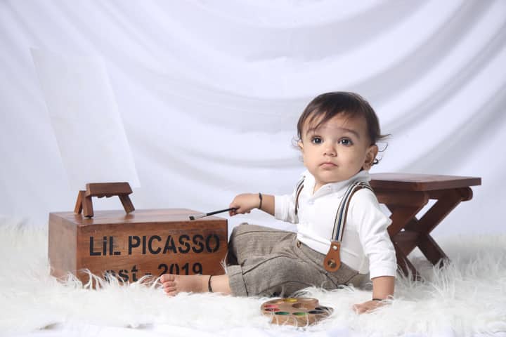 Adorable baby dressed as an artist playing with a paintbrush and easel in a cozy setting.