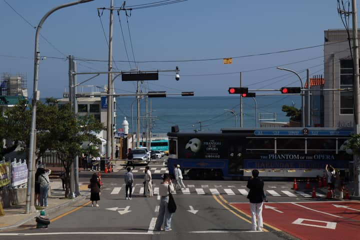 People watching a tourist train crossing near a coastal road with ocean in the background.
