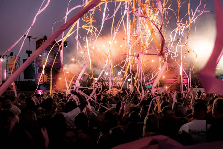 Crowd enjoying a vibrant festival with colorful ribbons and stage lights under a twilight sky.
