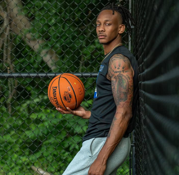 Athletic adult man holding basketball against a fence outdoors, showcasing tattoos and sports attire.
