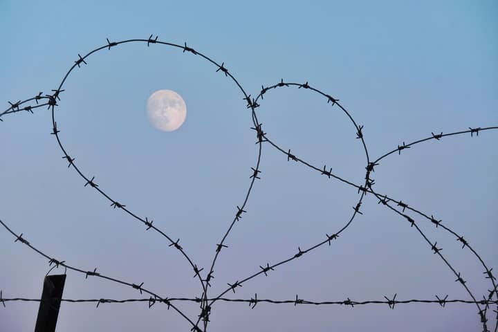Barbed wire forms heart shapes with the moon in a clear blue sky.
