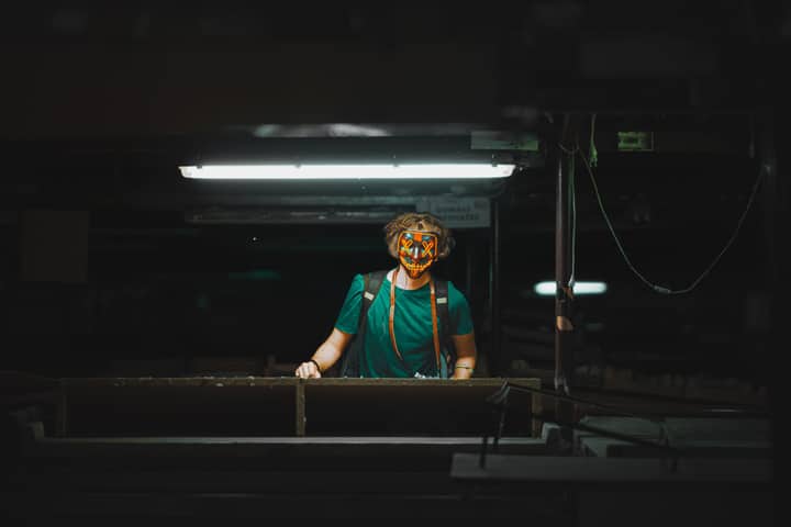 A masked person stands under a fluorescent light in an industrial environment, creating a mysterious and moody atmosphere.