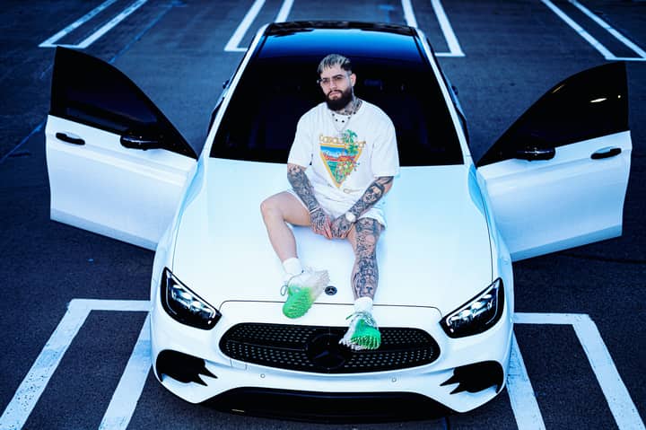 Tattooed man sitting on a white luxury car with open doors in an urban parking lot.