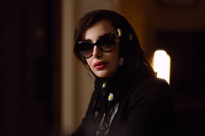 Elegant portrait of a woman wearing sunglasses and a floral shawl in a dimly lit room.