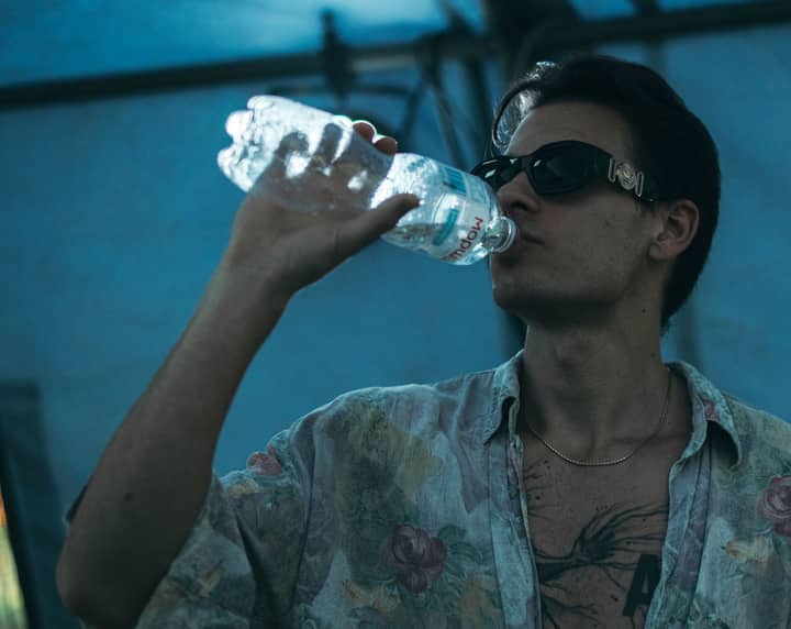 Casually dressed man sipping water from a bottle under a blue canopy.