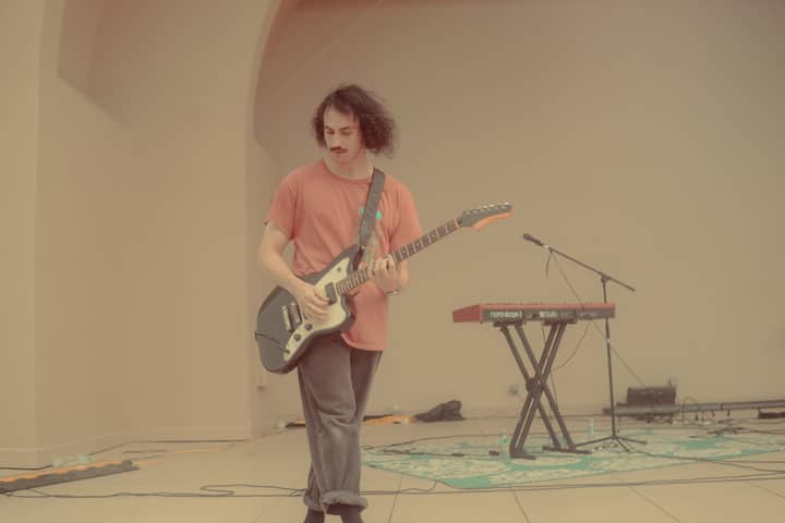 A young musician with a mustache playing an electric guitar on stage indoors.