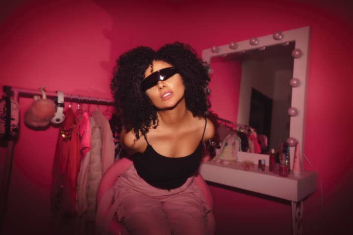Woman with curly hair and sunglasses posing stylishly in a vibrant pink room.