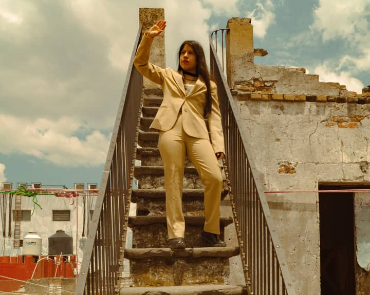Woman in a beige suit standing on a rooftop staircase against a rustic urban backdrop.