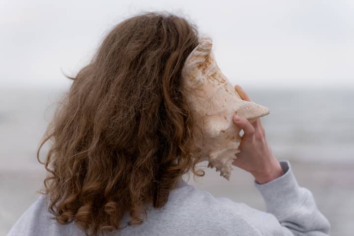 Rear view of a woman holding a seashell to her ear by the ocean.