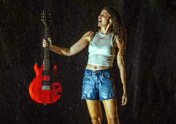 A woman stands boldly in the rain, holding a red electric guitar, under nighttime lighting.