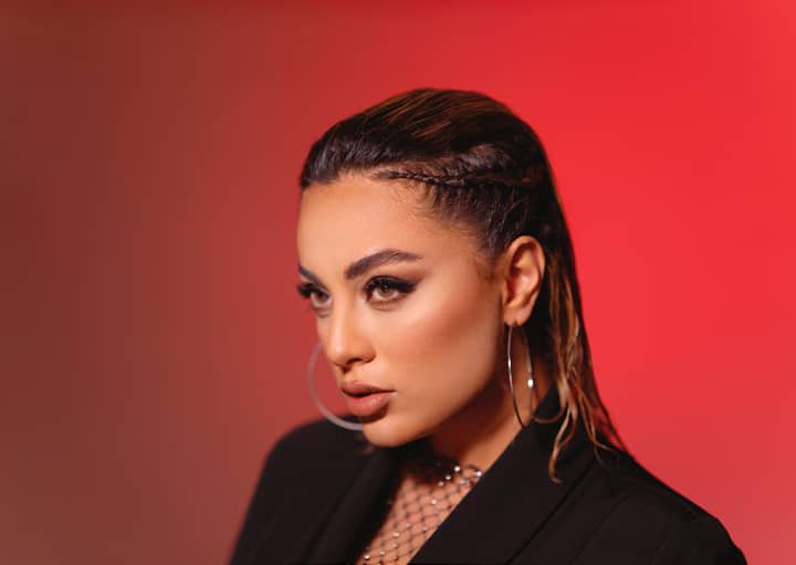 Close-up portrait of a woman with braided hair and hoop earrings against a red background.