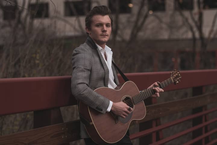 A man playing acoustic guitar on a bridge with a stylish outfit.
