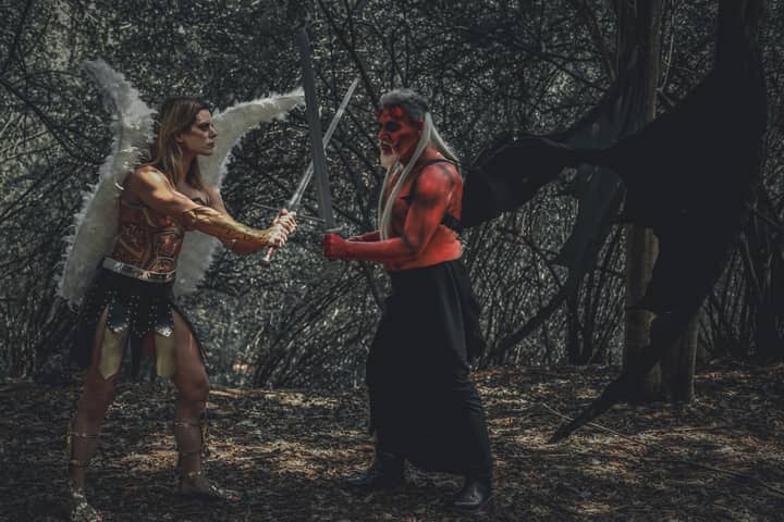 A dramatic cosplay scene of an angel and demon with swords in a forest.