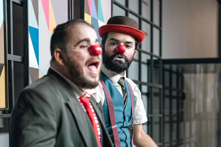 Two male clowns with red noses perform expressively on stage, adding humor and charm.