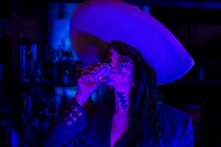 A woman with elegant Catrina makeup drinks in neon blue light, celebrating Mexican culture.