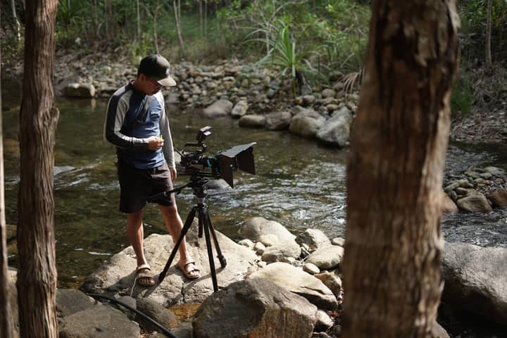 A cameraman operates equipment beside a stream in a forest setting in Mati, Philippines.